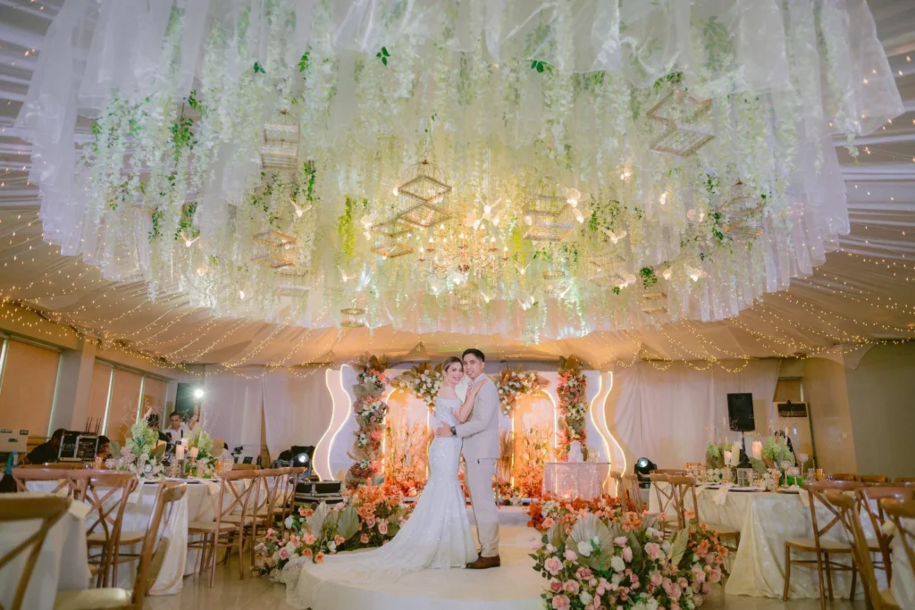 Bride and groom standing on an elegantly decorated wedding stage with floral and chandelier lighting.
